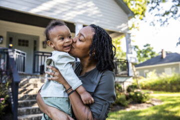 Black Mother holding and kissing happy toddler in front of new home